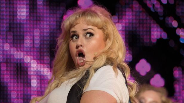 Rebel Wilson mit offenem Mund und überraschtem Gesichtsausdruck vor einem violetten Hintergrund.