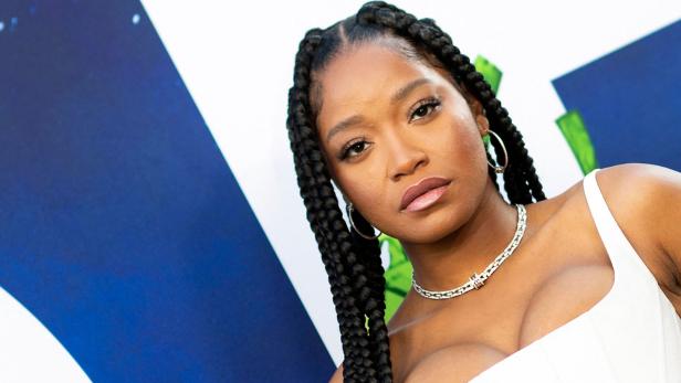 Keke Palmer posiert mit geflochtenen Zöpfen und weißem Oberteil vor einer Fotowand.