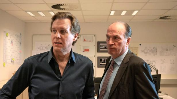 Zwei Männer stehen in einem Büro mit Plänen und Diagrammen an der Wand.