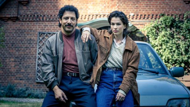 Ein Mann und eine Frau posieren lässig auf der Motorhaube eines Autos vor einem Backsteinhaus.