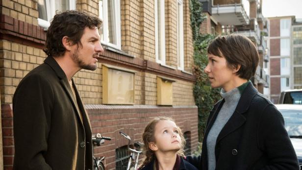 Ein Mann, eine Frau und ein junges Mädchen stehen vor einem Backsteingebäude.