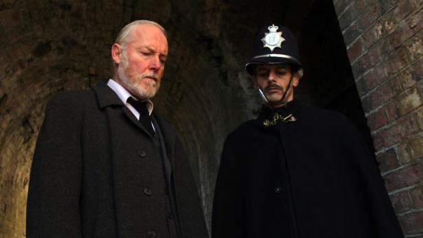 Ein Mann im Mantel und ein Polizist in historischer Uniform stehen in einem Tunnel.