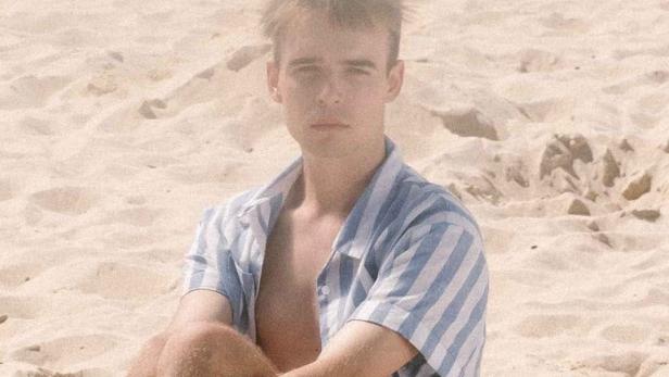 Ein junger Mann sitzt am Strand, im Hintergrund der Filmtitel „Reunion“.