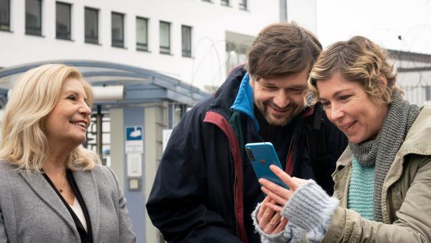 Drei Personen schauen sich ein blaues Smartphone vor einem Gebäude an.