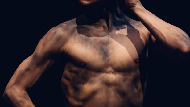 Ein Mann mit nacktem Oberkörper und schmutziger Haut posiert vor einem dunklen Hintergrund.