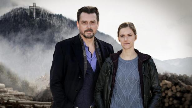 Ein Mann und eine Frau stehen vor einer Berglandschaft mit einem Kreuz auf einem Hügel.