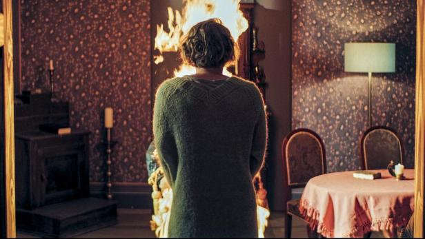 Eine Frau steht vor einem brennenden Kamin in einem altmodischen Zimmer.