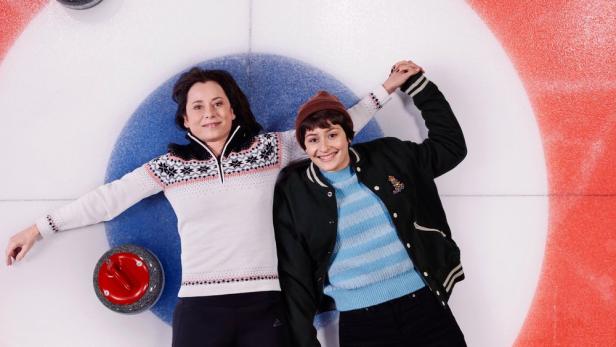 Zwei Frauen liegen auf einer Eisbahn mit Curling-Zielscheibe und halten Händchen.