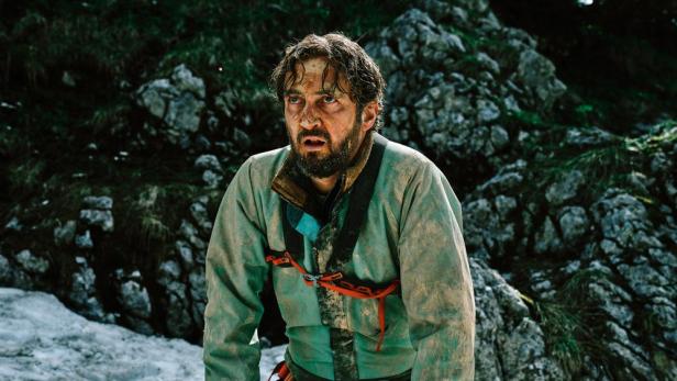 Ein verschmutzter Mann mit Klettergurt steht vor einem felsigen Hintergrund.