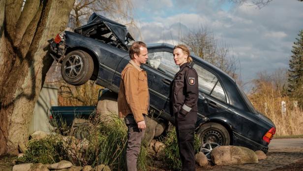 Ein Mann und eine Polizistin stehen vor einem zerstörten Auto, das auf einem Baumstumpf liegt.