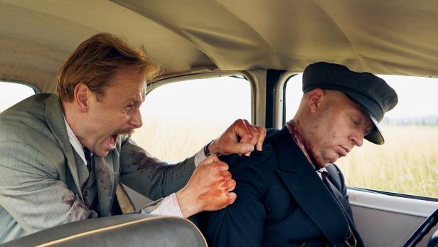 Ein Mann im Anzug schlägt einen bewusstlosen Mann mit Mütze im Auto.