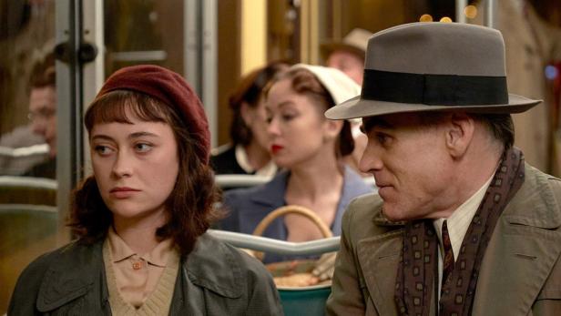 Eine junge Frau mit roter Baskenmütze und ein älterer Mann mit Hut sitzen in einem Zug.