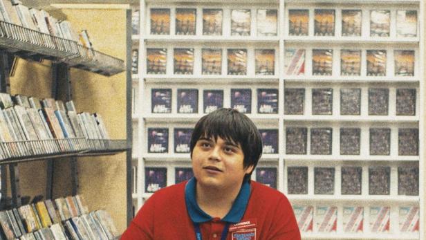 Ein junger Mann sitzt in einem Videoverleih vor Regalen voller Filme.