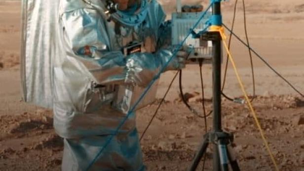 Ein Astronaut in einem Raumanzug arbeitet an einer Antenne in einer Wüstenlandschaft.