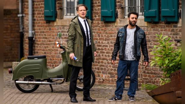 Zwei Männer stehen vor einer roten Backsteinmauer und einem olivgrünen Motorroller.