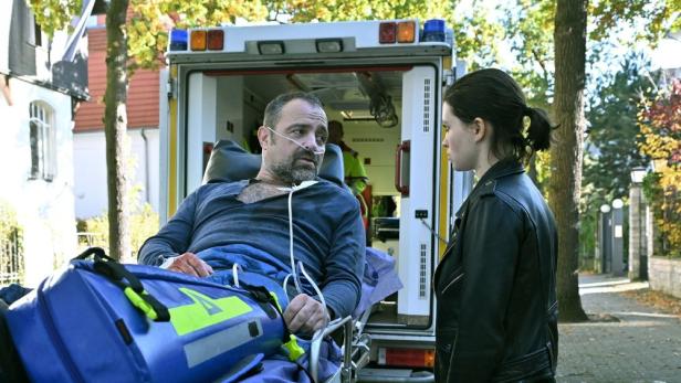 Ein Mann liegt mit einer Sauerstoffmaske auf einer Trage vor einem Krankenwagen, während eine Frau mit ihm spricht.