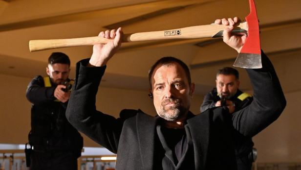 Ein Mann hält eine Axt über den Kopf, während zwei Polizisten im Hintergrund ihre Waffen ziehen.