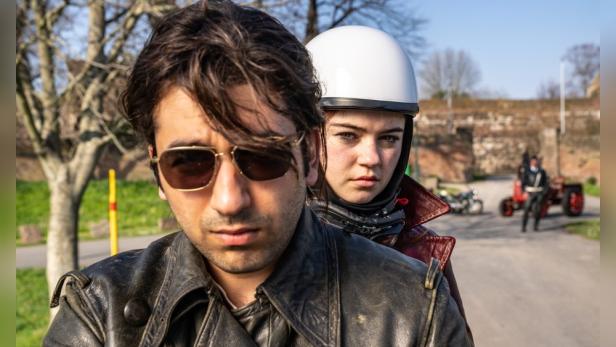 Ein Mann mit Sonnenbrille und eine Frau mit Motorradhelm sitzen auf einem Motorrad.