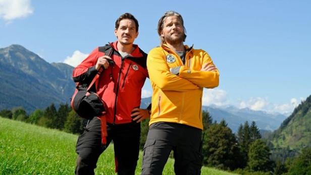 Zwei Schauspieler der Show "Die Bergretter" in einer roten und gelben Jacke stehen vor einer grünen Wiese.