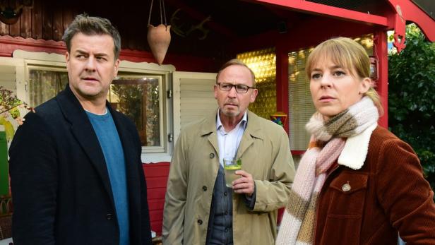 Drei Personen stehen vor einem rot gestrichenen Haus, möglicherweise für die Serie „München Mord“.