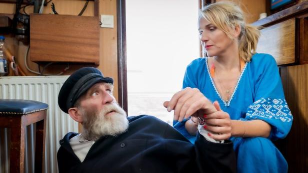 Eine Frau hält die Hand eines älteren Mannes mit Bart und Kapitänsmütze.