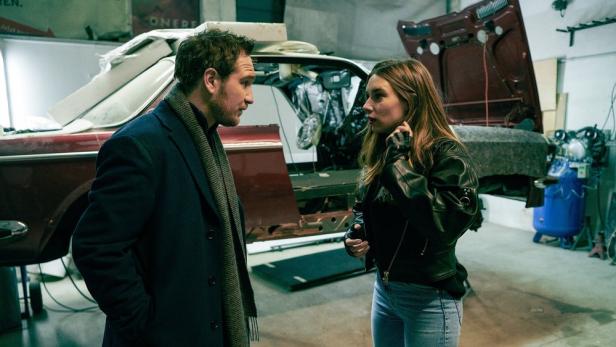Ein Mann und eine Frau stehen in einer Werkstatt vor einem Oldtimer und diskutieren.