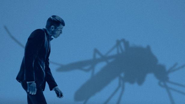 Ein Mann in Anzug geht unter dem Schatten einer riesigen Mücke entlang, Titel „Mosquito State“ im Hintergrund.