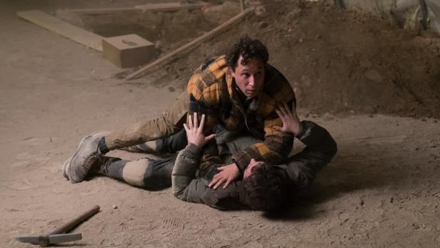 Zwei Männer ringen auf einem staubigen Boden, ein Pickel liegt daneben.