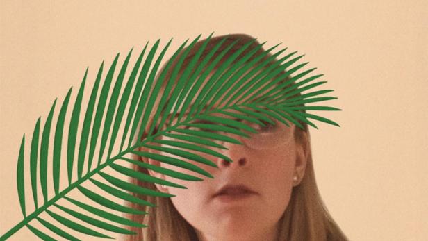 Eine Frau mit blonden Haaren, teilweise von einem grünen Palmblatt bedeckt, vor beigem Hintergrund.