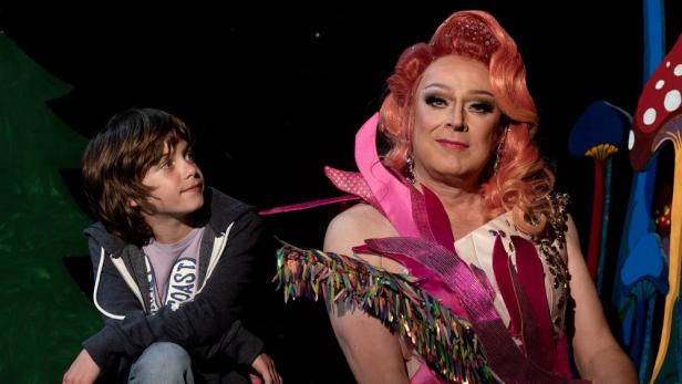 Ein Kind blickt zu einer Dragqueen mit orangefarbenem Haar und einem pinkfarbenen Kleid auf einer Bühne.