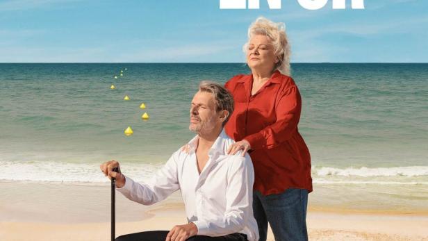 Ein Mann sitzt mit einem Stock am Strand, während eine Frau hinter ihm steht.