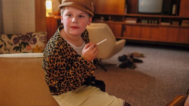 Ein Junge im Leopardenmuster-Jacket und Hut sitzt mit einer Zigarette in der Hand.