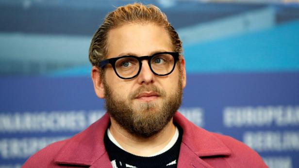 Porträt von Jonah Hill mit Brille und rosa Jacke vor blauem Hintergrund.