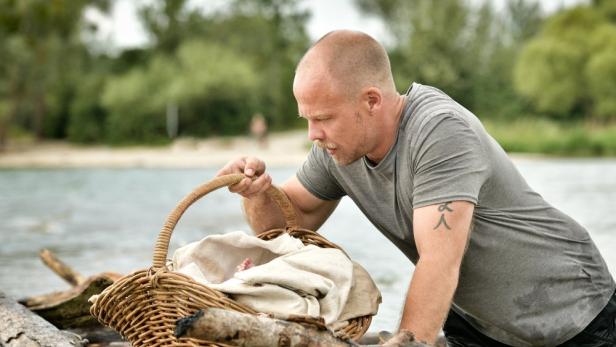 Ein Mann beugt sich über einen Korb mit einem Baby am Ufer eines Sees.