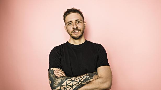 Ein Mann mit Tätowierung auf dem Arm lächelt vor einem rosa Hintergrund.