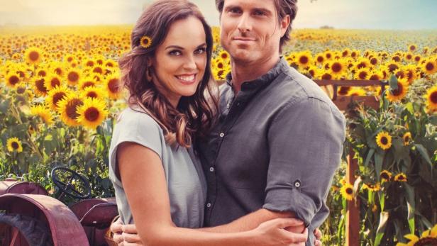 Ein Paar umarmt sich vor einem Feld voller Sonnenblumen, im Hintergrund ein alter Traktor.