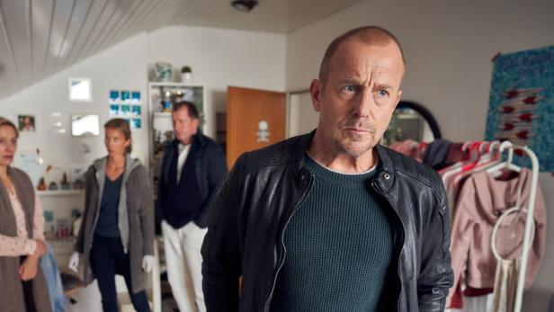 Ein Mann in Lederjacke steht in einem Raum, während im Hintergrund weitere Personen zu sehen sind.