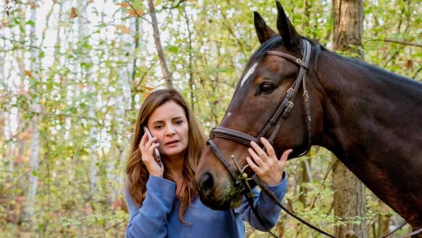 Eine Frau telefoniert besorgt neben einem braunen Pferd im Wald.