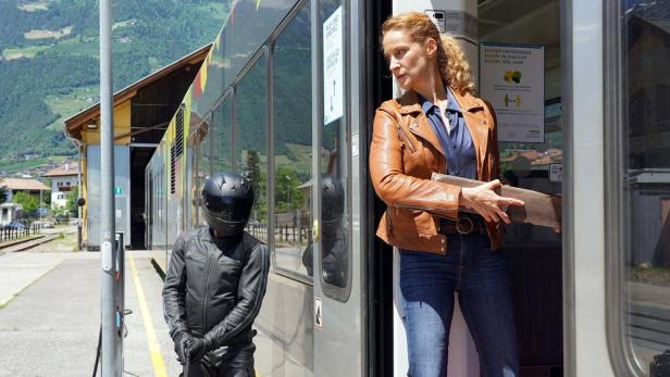 Eine Frau mit einer Holzplanke steigt aus einem Zug, während ein Motorradfahrer wartet.