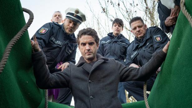 Ein Mann in einem grünen Behältnis wird von mehreren Polizisten umringt.