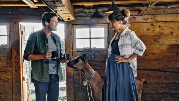 Ein Mann bringt einer Frau in einem Holzhaus Kaffee, während eine Ziege zusieht.
