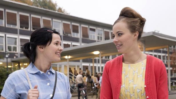 Zwei junge Frauen im Stil der 1960er Jahre unterhalten sich vor einem Schulgebäude.