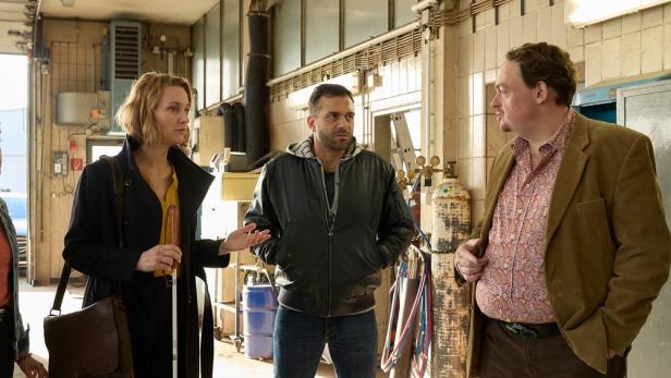 Drei Personen, darunter eine blinde Frau mit Stock, unterhalten sich in einer Werkstatt.