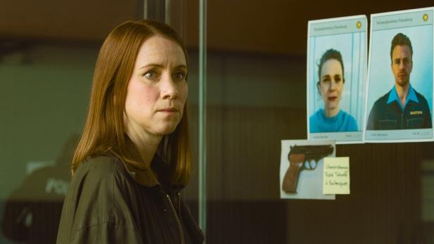 Eine Frau steht vor einer Wand mit Fotos von Verdächtigen und einer Waffe.