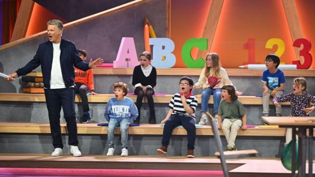 Ein Moderator steht vor einer Gruppe aufgeregter Kinder vor einer bunten Schulumgebung für die Sendung „Zurück in die Schule“.