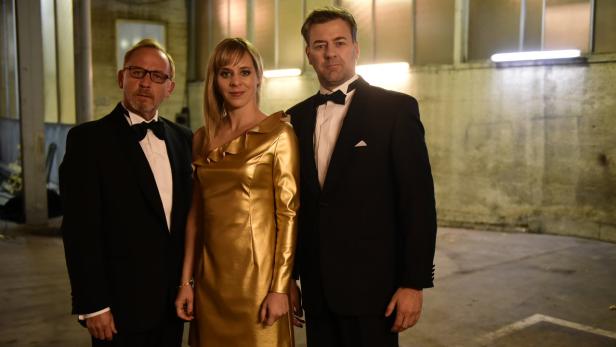Zwei Männer im Smoking und eine Frau in einem goldenen Kleid posieren für ein Foto.