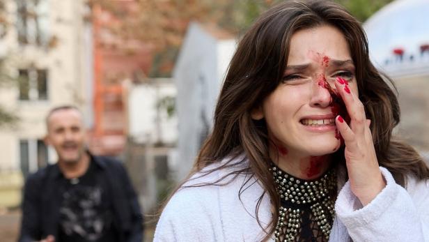 Eine weinende Frau mit blutverschmiertem Gesicht, während ein Mann im Hintergrund grinst.