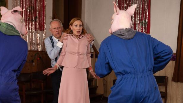 Eine verängstigte Frau in Rosa wird von einem Mann beschützt, während zwei Personen mit Schweinsmasken bedrohlich wirken.