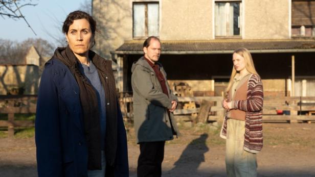 Eine Frau steht besorgt vor einem Haus, während ein Mann und ein junges Mädchen im Hintergrund stehen.