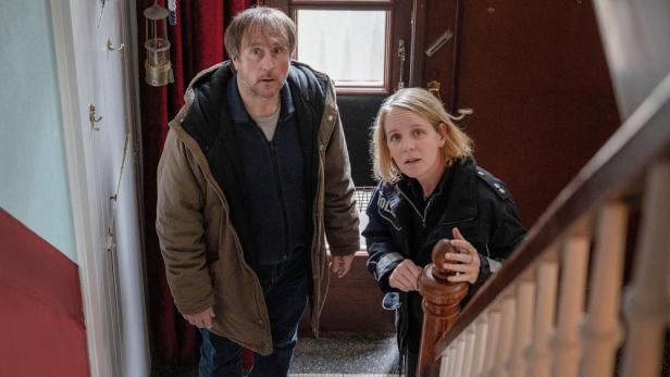 Ein Mann und eine Polizistin stehen überrascht in einem Treppenhaus.